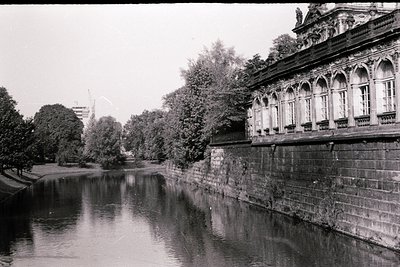 Neoclassical riverside building with arched colonnades and balustrades, reflecting in calm waters. Lush greenery frames the s...
