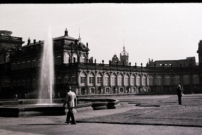 Neoclassical palace courtyard with grand arched colonnade, central fountain, and symmetrical design. Two men in mid-20th-cent...
