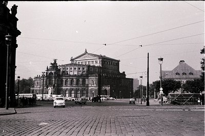 Grand neoclassical building with ornate pediment and columns dominates a mid-20th-century European plaza. Cobblestone streets...