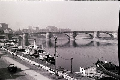 Black-and-white urban riverscape featuring a multi-arched stone bridge spanning a wide river. Industrial-era barge traffic do...