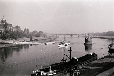 Black-and-white river scene featuring a stone bridge spanning a wide waterway. Historic European architecture with spires and...