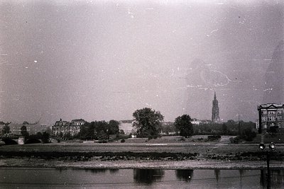 Vintage black-and-white cityscape featuring a prominent church spire and riverside buildings, likely European. Dense foliage ...