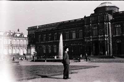 Grand Baroque-style palace courtyard with ornate façade, arched windows, and domed towers. A man in 1960s-era suit poses near...