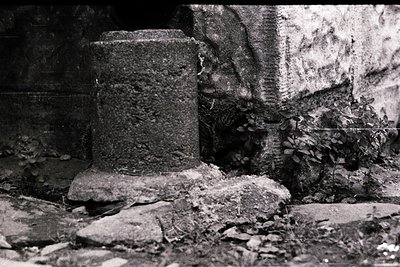 Weathered concrete column base and stone foundation in overgrown ruins, suggesting abandoned infrastructure. Textured erosion...