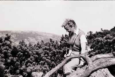 Black-and-white portrait of a person sharpening a knife on a rocky outcrop, surrounded by dense pine forest. Mid-20th century...