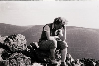 Mid-20th century black-and-white photo of a lone individual resting atop rugged rock formations, hands resting on knees. Stri...