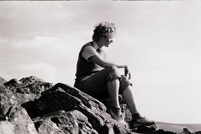 Mid-century outdoor portrait of a person in casual hiking attire—short-sleeve shirt, rolled pants, and sturdy boots—sitting o...