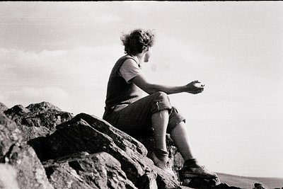 Mid-century outdoor portrait of a person sitting on rugged rocks, holding a small object. Classic 1960s/70s composition with ...