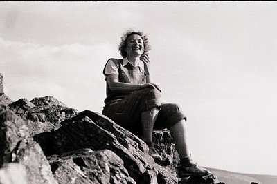 Black-and-white portrait of a woman seated on rugged rocks, smiling at the camera. She wears a striped blouse, pleated skirt,...