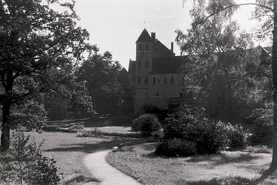 Mid-20th century castle-like villa with steep gabled roof and tower, nestled in a lush, winding garden path. Stone walls and ...