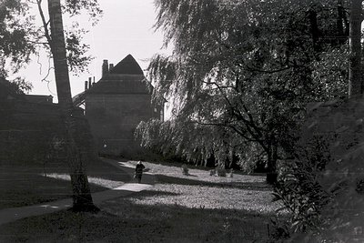Black-and-white shot of a lone figure walking along a gravel path in a sunlit park. Distinctive stone building with steep gab...