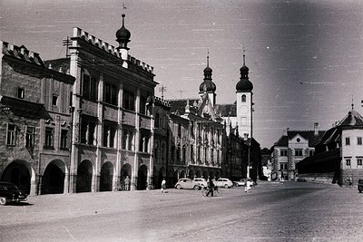 Black-and-white street scene featuring **1950s-era European architecture**—prominent **arcaded buildings** with classical fac...