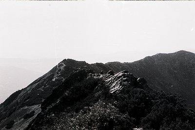 Black-and-white aerial view of rugged volcanic terrain with jagged rock formations and sparse vegetation. The layered, eroded...