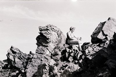 Mid-century black-and-white photo of a woman seated atop jagged volcanic rock formations, likely in a coastal or island setti...