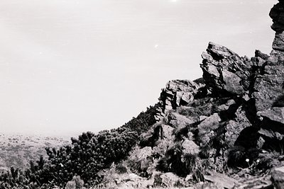 High-angle black-and-white shot of rugged, rocky terrain with sparse vegetation. Dramatic cliffs frame a distant, hazy valley...