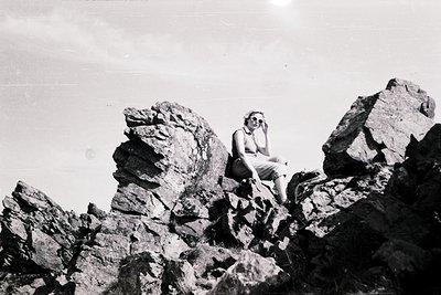 Mid-century outdoor portrait of a woman seated on rugged rock formations, holding a camera. Classic 1950s–1960s travel photog...
