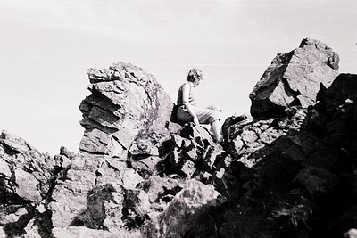 A lone individual in mid-20th-century attire climbs jagged rock formations, suggesting rugged outdoor activity. The black-and...