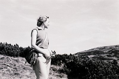 Mid-century woman in a sleeveless ribbed top and high-waisted trousers stands on a grassy hillside, clutching a small bouquet...