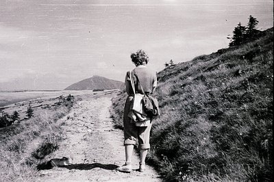 Mid-20th century hiker on a rugged coastal trail, back to camera, wearing a striped sweater and backpack. Dramatic seascape w...