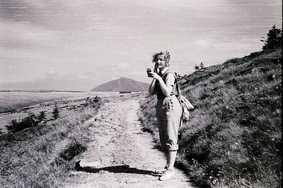 Mid-20th century hiker pauses on rugged coastal trail, holding a vintage camera. Volcanic peak and ocean backdrop suggest a r...