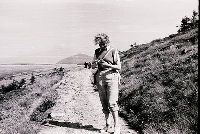 Mid-20th century black-and-white photo of a woman hiking on a rugged coastal trail, holding a map or booklet. She wears a swe...