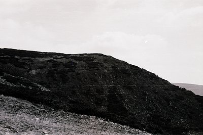 Black-and-white shot of rugged alpine terrain with steep rock face and sparse vegetation. Single climber in mid-ascent on rig...
