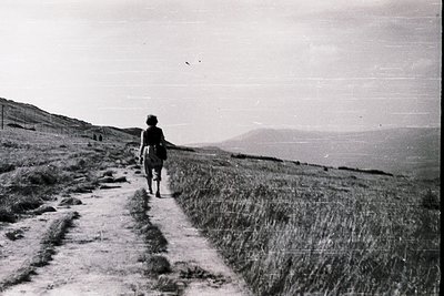 Mid-20th century black-and-white photo of lone hiker on a rural dirt path, framed by grassy hills and distant mountains. The ...