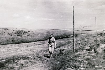 Man in military uniform walks along a barren, fenced-off path, likely a prisoner or guard in a mid-20th-century detention or ...