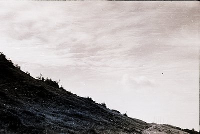 Vintage black-and-white aerial view of terraced hillside with sparse vegetation, likely agricultural. Clouds obscure mid-sky,...