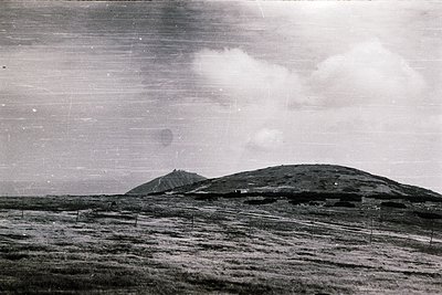 Vintage black-and-white landscape of a barren, grassy hillside with scattered fencing and a lone structure near the base. Dra...