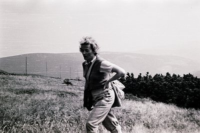 Black-and-white photo of a man mid-stride on a grassy hillside, carrying a folded jacket and backpack. Rolling hills and spar...