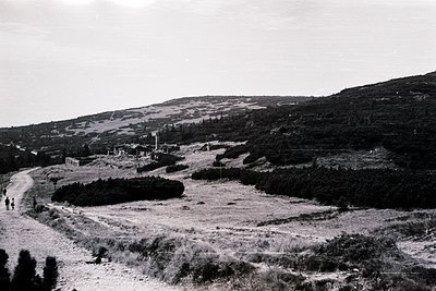 Black-and-white landscape featuring a winding dirt road through a rural, hilly terrain. A lone figure walks ahead, while scat...