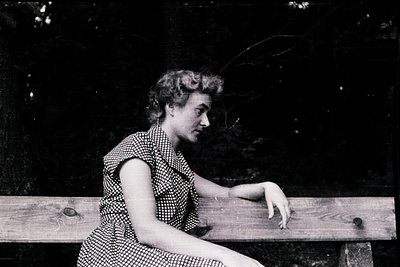 Black-and-white portrait of a woman in a polka-dot dress, seated on a wooden bench outdoors. Her posture suggests contemplati...