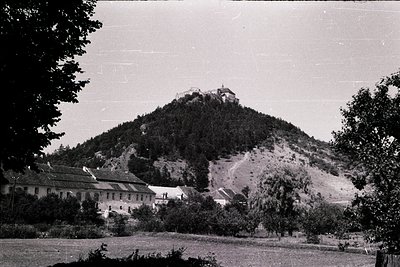 Vintage black-and-white shot of a hilltop fortress atop rugged terrain, surrounded by dense forest. Foreground features a low...