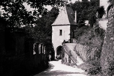 Historic stone gatehouse with steep gabled roof, flanked by ivy-covered walls. Mid-20th century European architectural style....