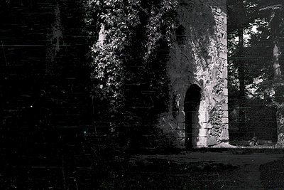 Vintage black-and-white shot of a weathered stone archway flanked by aged brick walls, partially obscured by foliage. Archite...