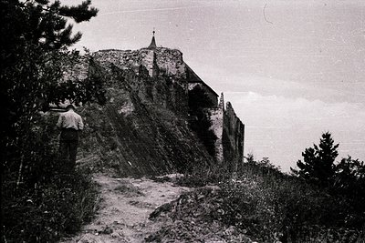 Black-and-white shot of a lone figure ascending a rocky path toward a medieval stone church perched on a cliffside. The churc...