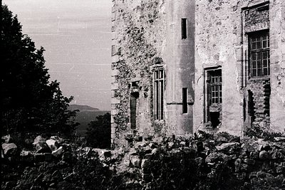 Abandoned stone building with weathered walls and broken windows, surrounded by overgrown vegetation. Architectural details i...