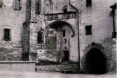 Historic stone façade featuring arched doorway and balcony with worn plaster. Architectural details include a small arched wi...