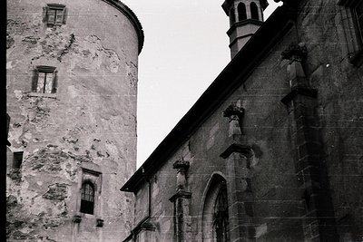 Gothic-style stone tower with weathered facade and small arched windows, adjacent to a building featuring a pointed arch door...