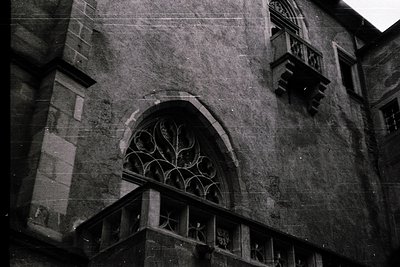 Gothic-style stone arch with intricate tracery window, flanked by weathered walls and wrought-iron balcony. Likely European m...