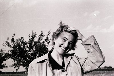 Mid-century woman in a relaxed pose, hand resting on shoulder, wearing a collared jacket and checkered blouse. Outdoor settin...