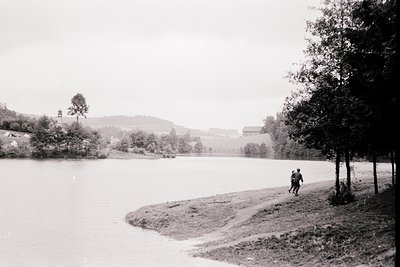 Black-and-white lakeside scene with lone figure walking along shoreline. Distant church steeple and rural buildings frame ser...