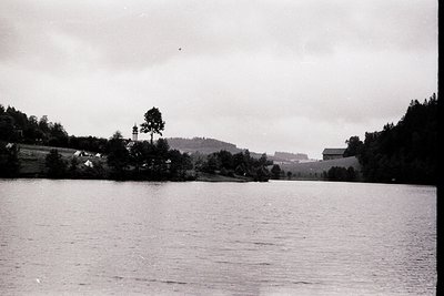 Black-and-white lakeside scene with dense forest framing a calm body of water. Distant farmhouses and rolling hills suggest r...