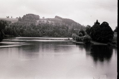 Mid-20th century black-and-white lakeside scene with dense forested hills framing calm waters. Small dock extends into lake, ...