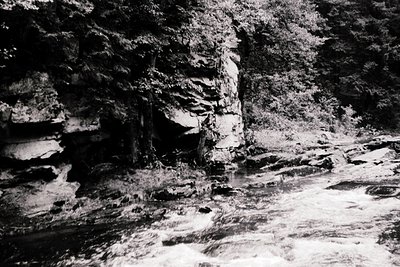 Black-and-white shot of a rocky riverbed with cascading water against vertical rock walls. Dense forest framing the scene sug...