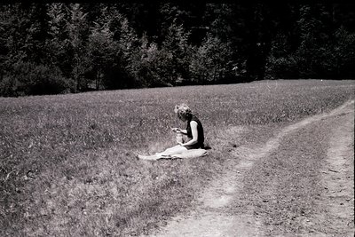 Black-and-white candid of a lone individual seated on a dirt path, engrossed in a small handheld device (likely a camera) in ...