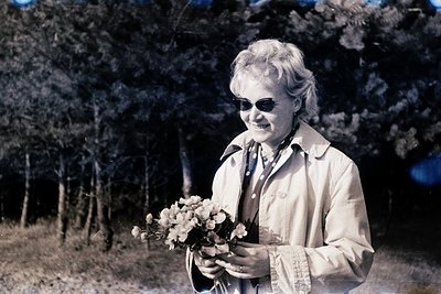 Vintage sepia-toned portrait of a woman in a light-colored coat, holding a bouquet of white flowers, outdoors in a wooded are...