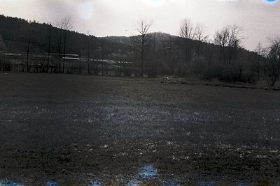 Black-and-white landscape featuring a muddy, flooded field with shallow water reflecting light. Bare trees and a distant rail...