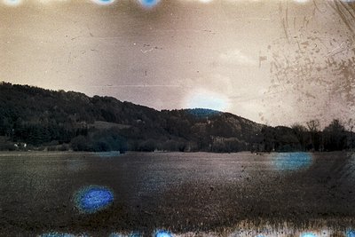 Vintage sepia-toned landscape featuring a serene lake bordered by forested hills. Distinctive lens flare and slight film grai...